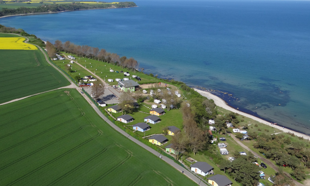 At KNAUS Campingpark Rügen, an overhead perspective reveals a campsite featuring cabins and recreational vehicles situated along a shoreline next to vibrant green meadows.