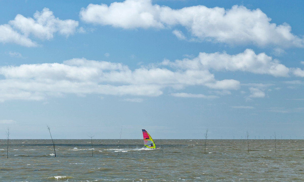 At KNAUS Campingpark Dorum, a windsurfer is skimming over a rough sea with dispersed tree markers beneath a partly cloudy sky.