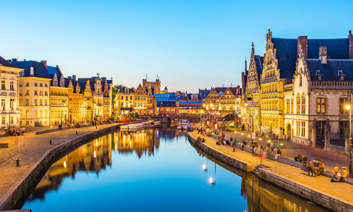 At Urban Gardens Gent, historic structures are mirrored in the canal at twilight, with individuals seated and strolling along the waterside.
