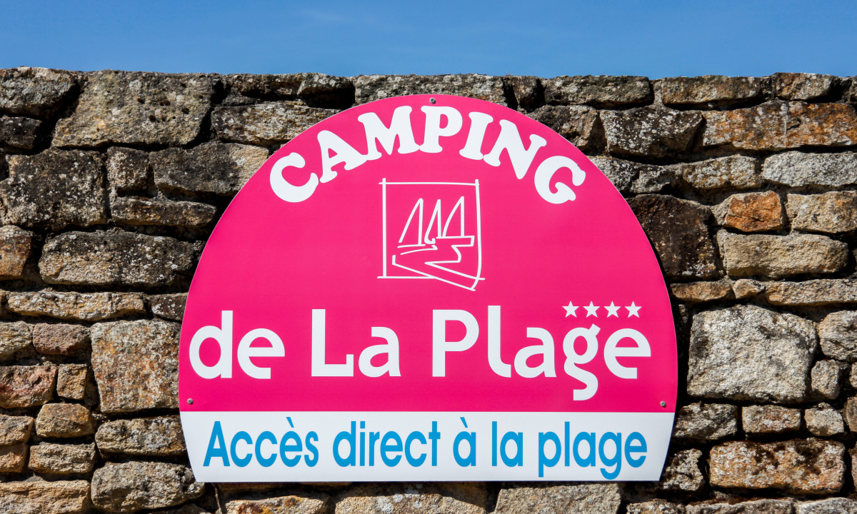At De la Plage, a pink circular sign is attached to a stone wall, featuring text and symbols, beneath a clear blue sky.