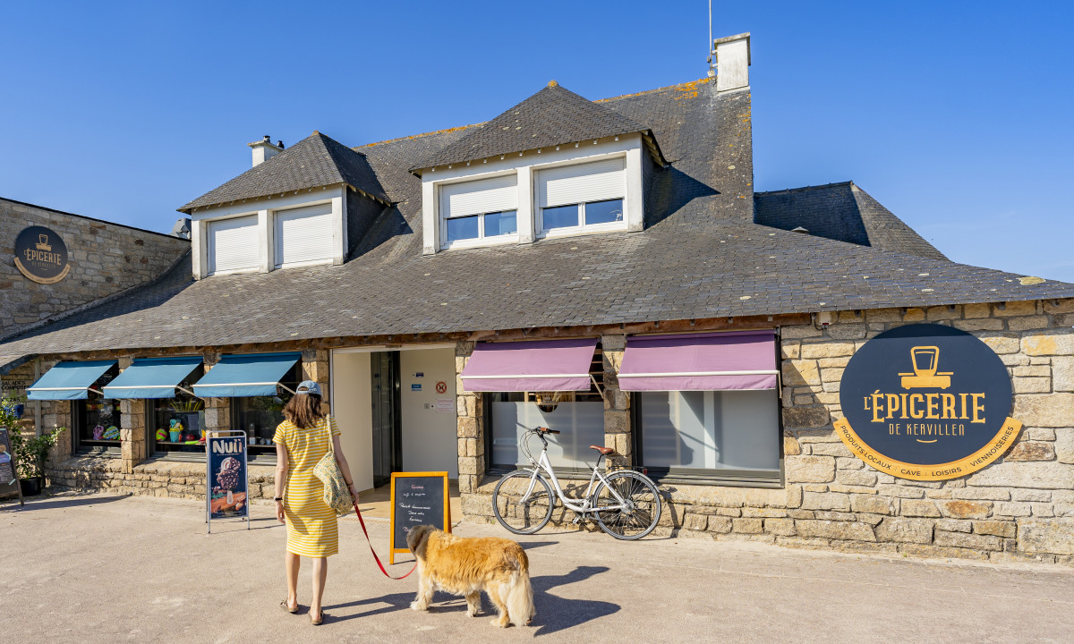 At De la Plage, a woman in a yellow gown strolls with her dog outside a stone shop, with bicycles secured nearby.