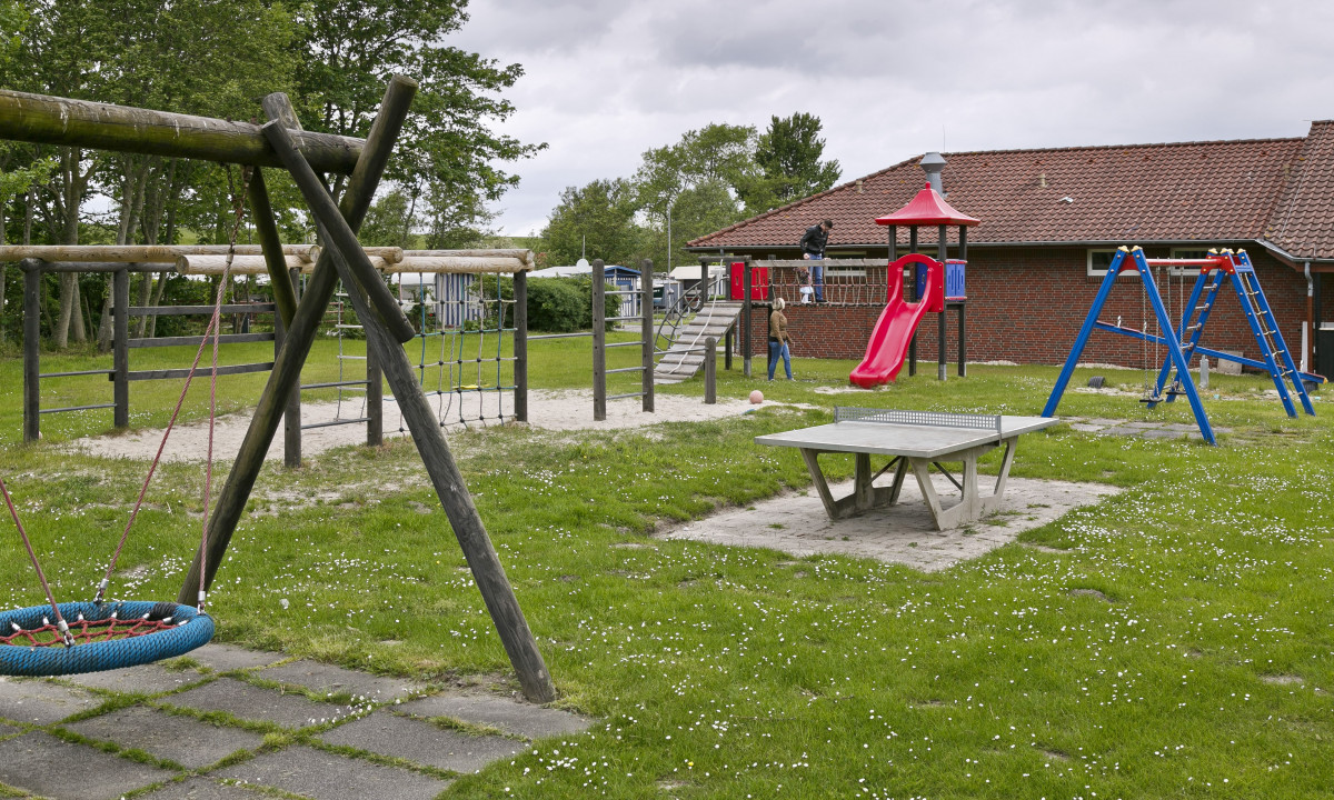 At KNAUS Campingpark Eckwarderhörne, there is a play area featuring swings, a slide, and climbing structures, all encircled by lush grass and trees adjacent to a brick structure.