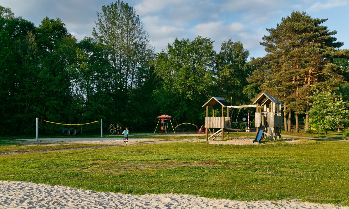 At KNAUS Campingpark Oyten, a wooden play structure featuring a slide is encircled by grass and trees, with a child running close by and a volleyball net visible in the background.