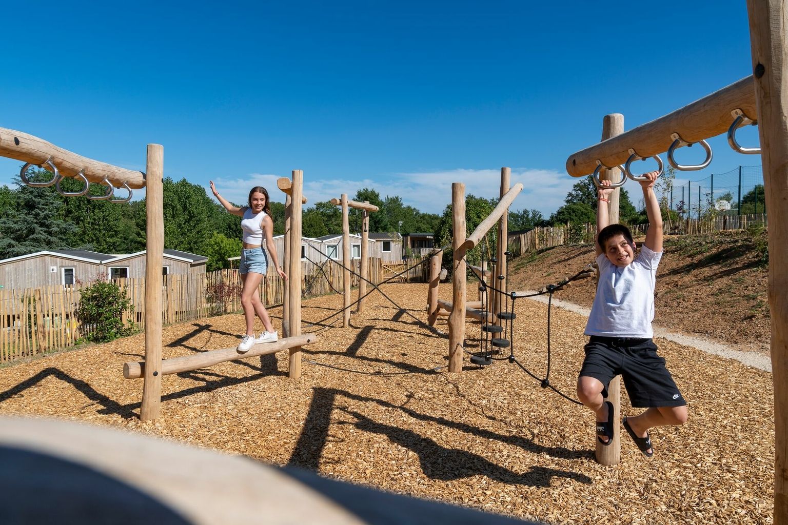 At Le Carbonnier, a child dangles from monkey bars while a woman maintains her balance on a beam in a playground, encircled by wooden cabins.