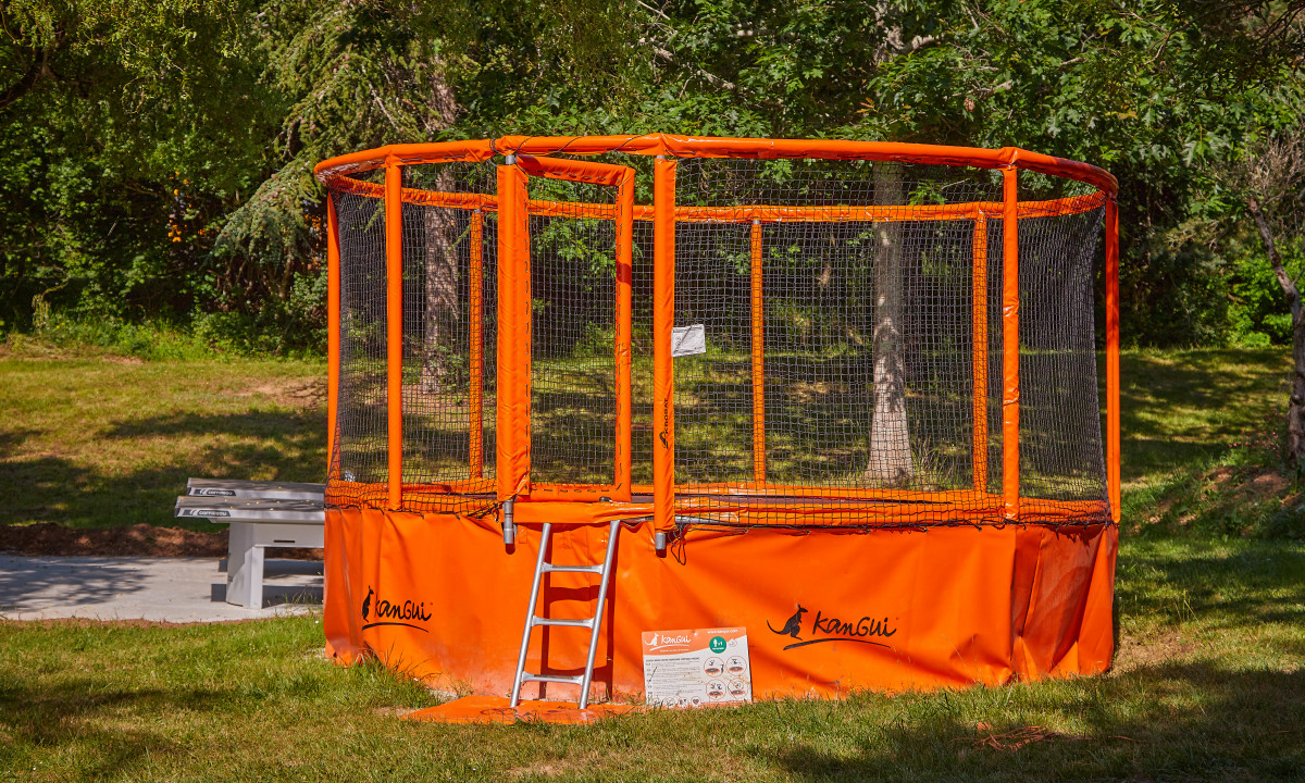 At Le Chêne Vert, a vibrant orange trampoline encircled by safety netting, accompanied by a small ladder, is situated in a grassy, wooded region.