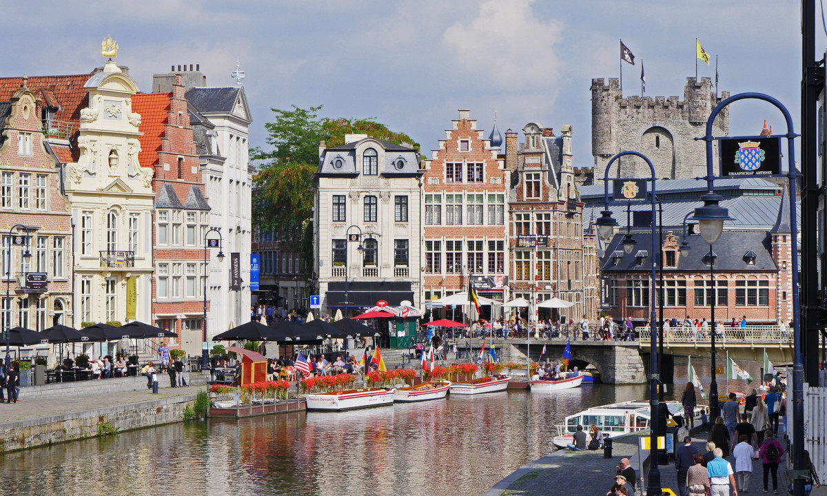 At Urban Gardens Gent, structures border the waterway where vessels are anchored; individuals stroll or relax at nearby cafes under a partially overcast sky.