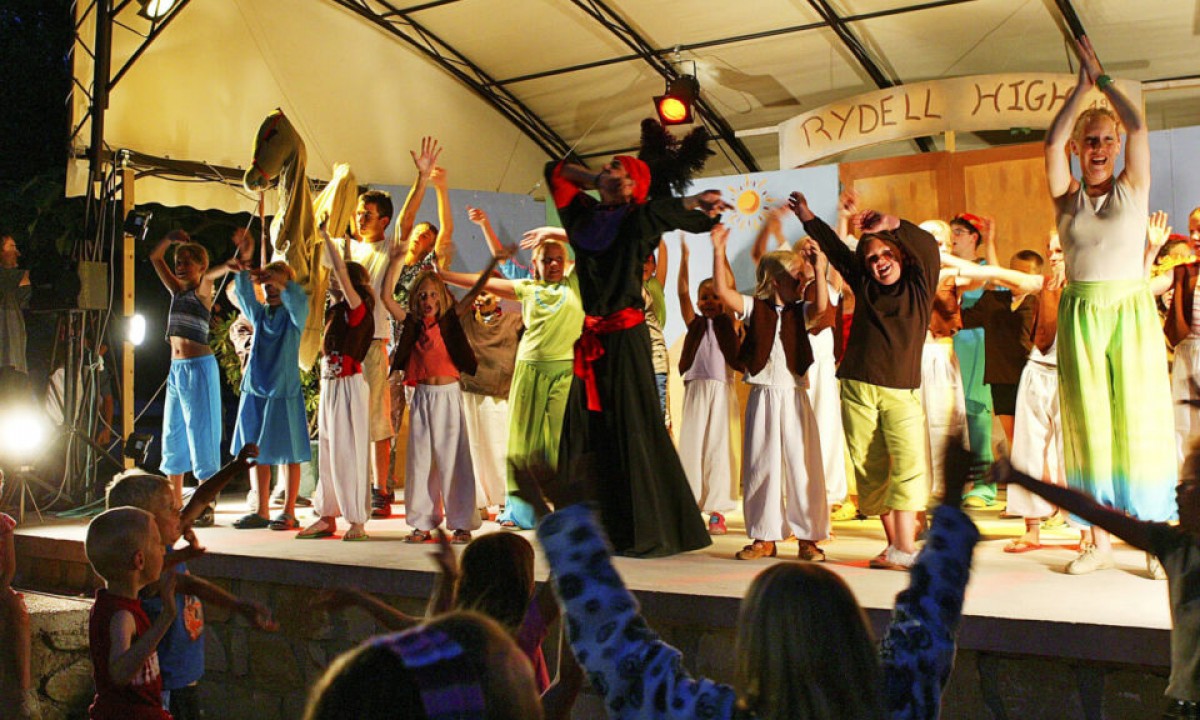 At Les Ranchisses, entertainers in outfits dance vigorously on a illuminated stage; spectators eagerly imitate the movements.