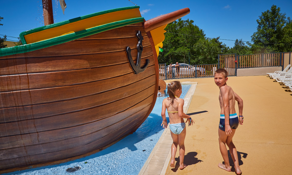At Terrasses du Lac, kids stroll around a pirate ship design in a shallow water play zone, accompanied by trees and lounge chairs in the vicinity.