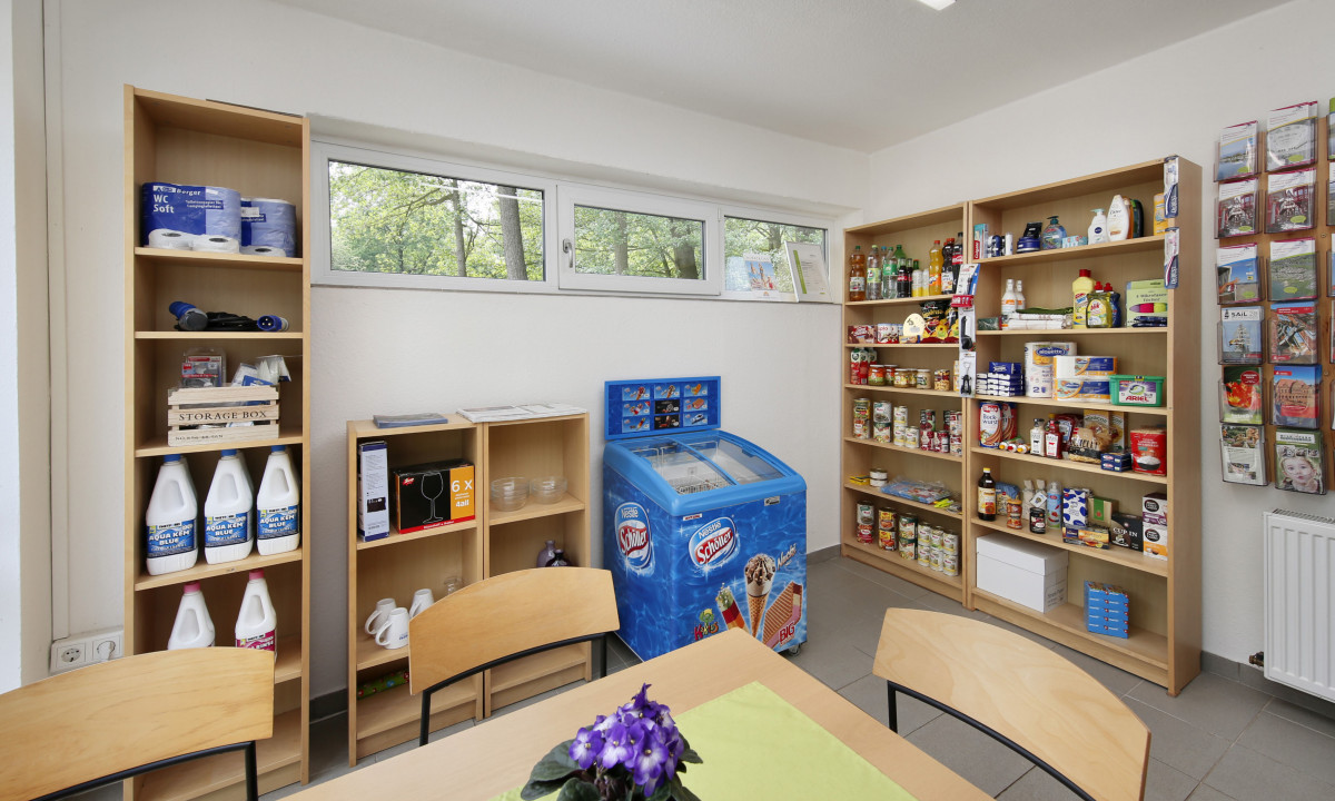 At KNAUS Campingpark Oyten, shelves containing cleaning products, food items, and an ice cream freezer are located in a small room adjacent to a table with chairs.