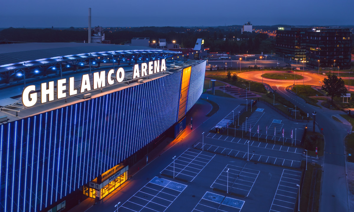 At Urban Gardens Gent, a spacious contemporary arena features a lit exterior during the evening, situated in an urban environment with adjacent parking areas.