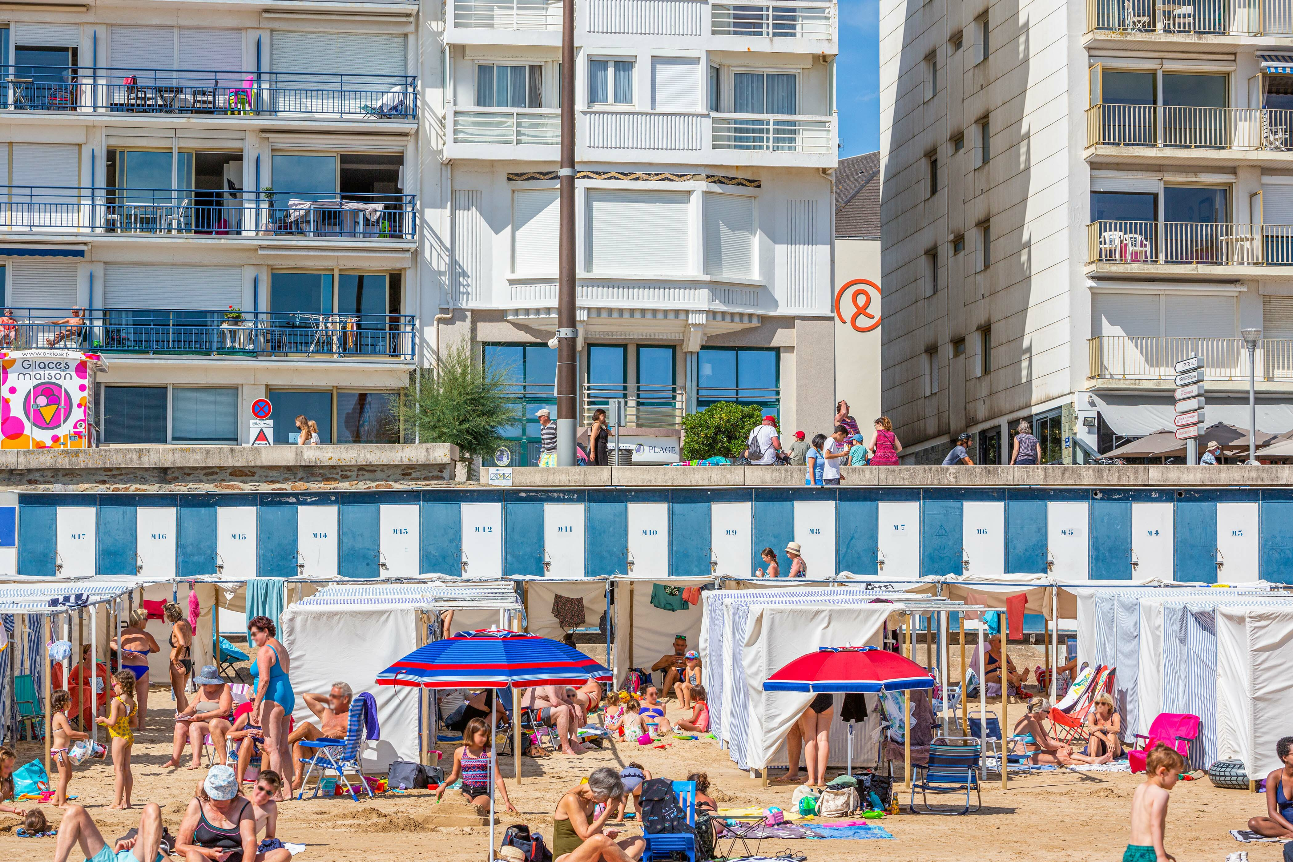 Beachgoers relaxing and socializing under umbrellas and tents near beachfront buildings at La Baie des Sables