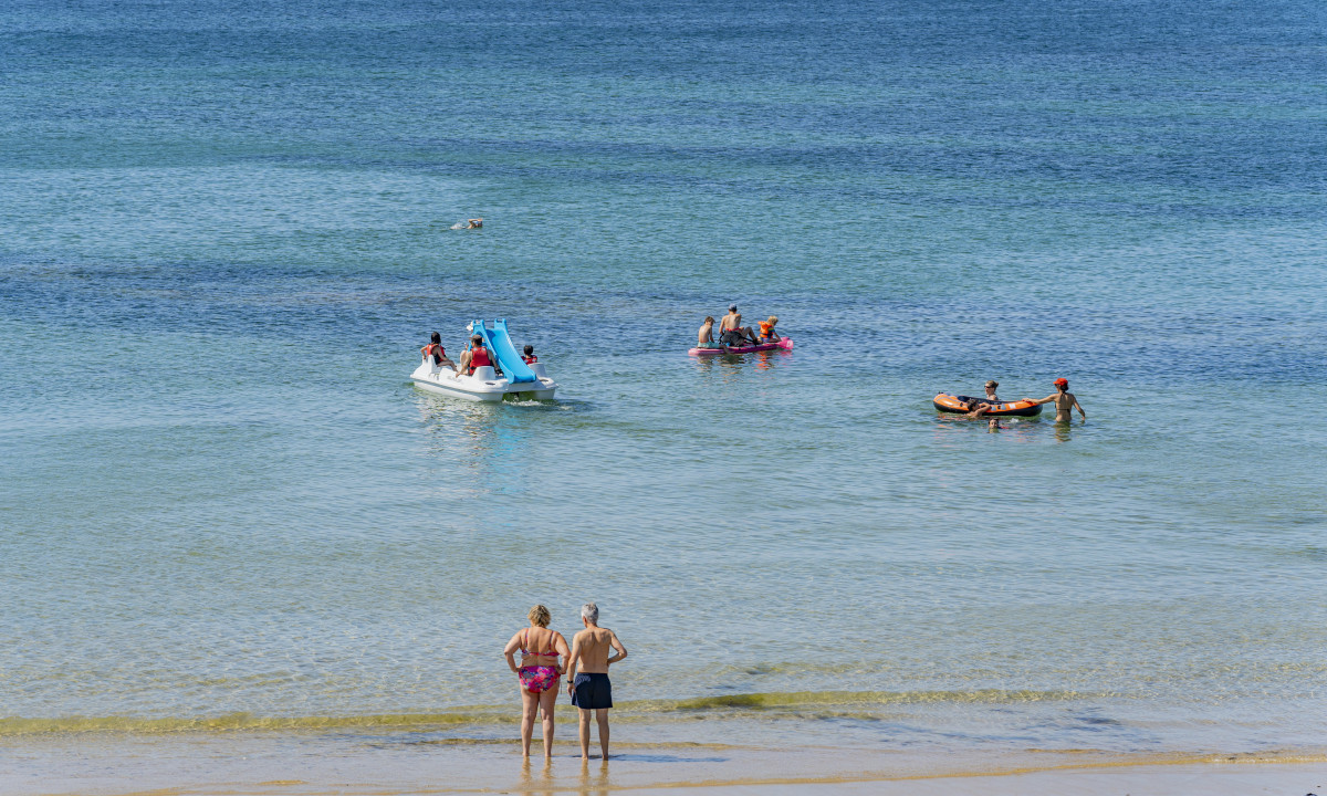 At De la Plage, individuals unwind on pedal boats and inflatable floats in tranquil, clear ocean waters alongside a sandy beach.