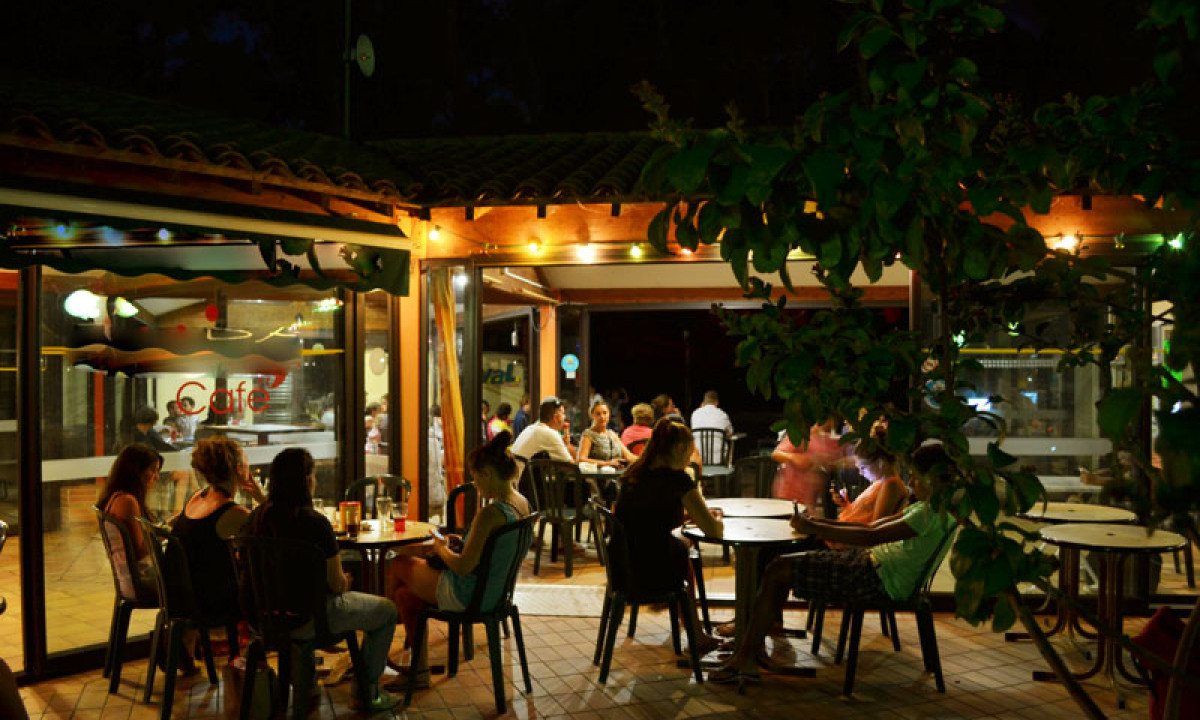At La Sousta, individuals are seated at outdoor café tables, savoring their evening under cozy lights, with the indoor space visible.