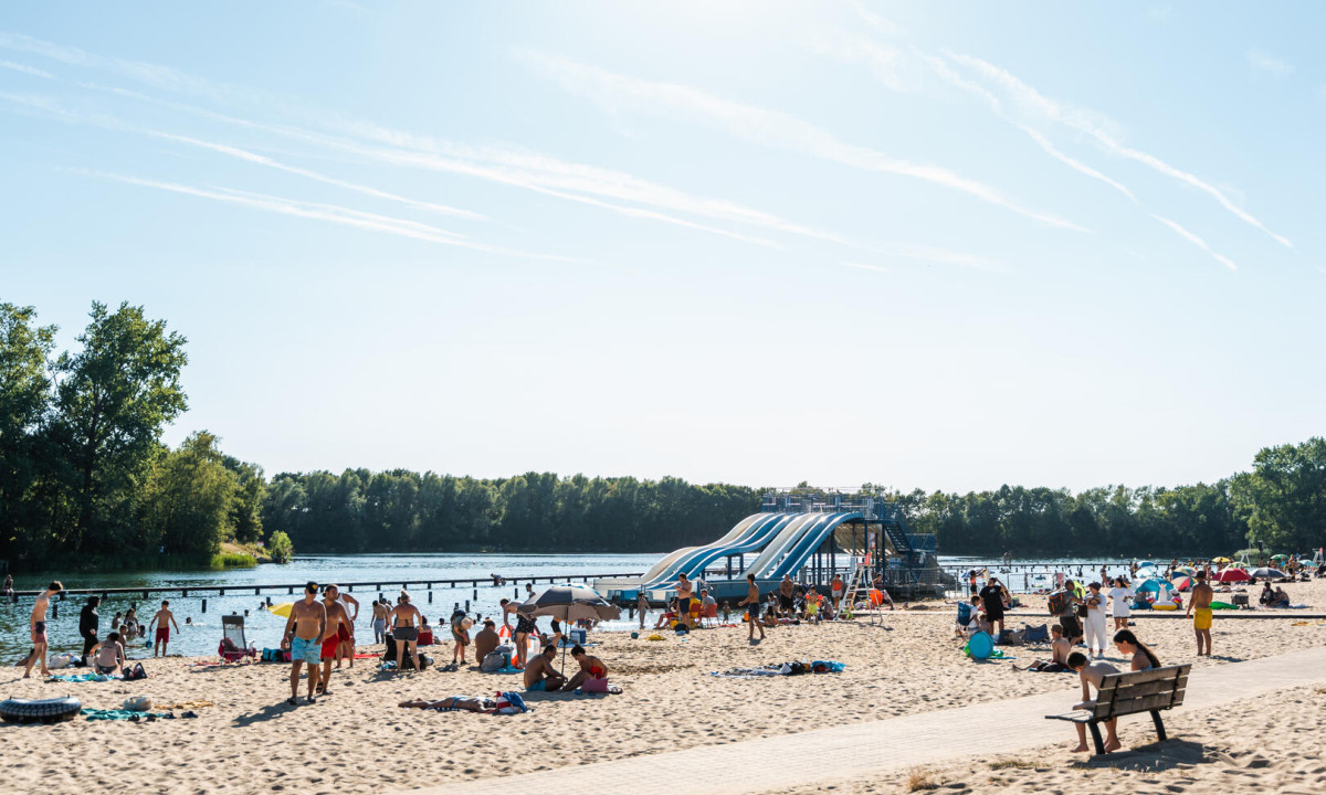 At Urban Gardens Gent, individuals are unwinding on a sandy shore with sizable water slides, encircled by trees and tranquil waters.