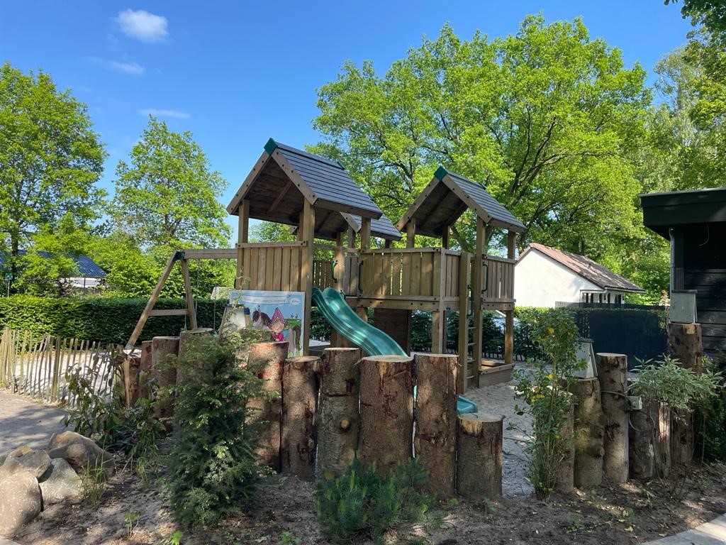 At de Vinkenkamp, there is a wooden play structure featuring slides and a climbing frame situated in a lush outdoor setting.