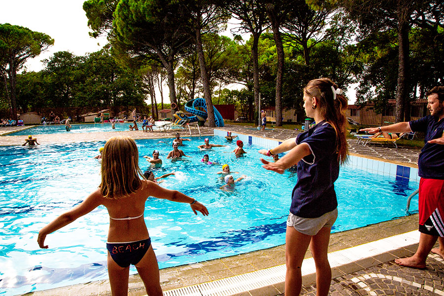 Children swimming and playing in a large, tree-surrounded pool while adults perform exercises at Belvedere Pineta