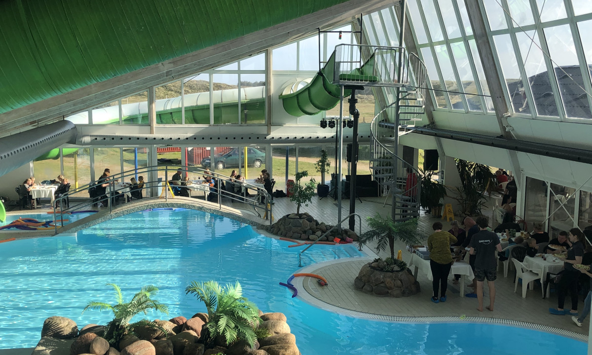 At Dancamps Nordsø, a swimming pool featuring a water slide is encircled by individuals enjoying meals at tables in a brightly lit indoor space.