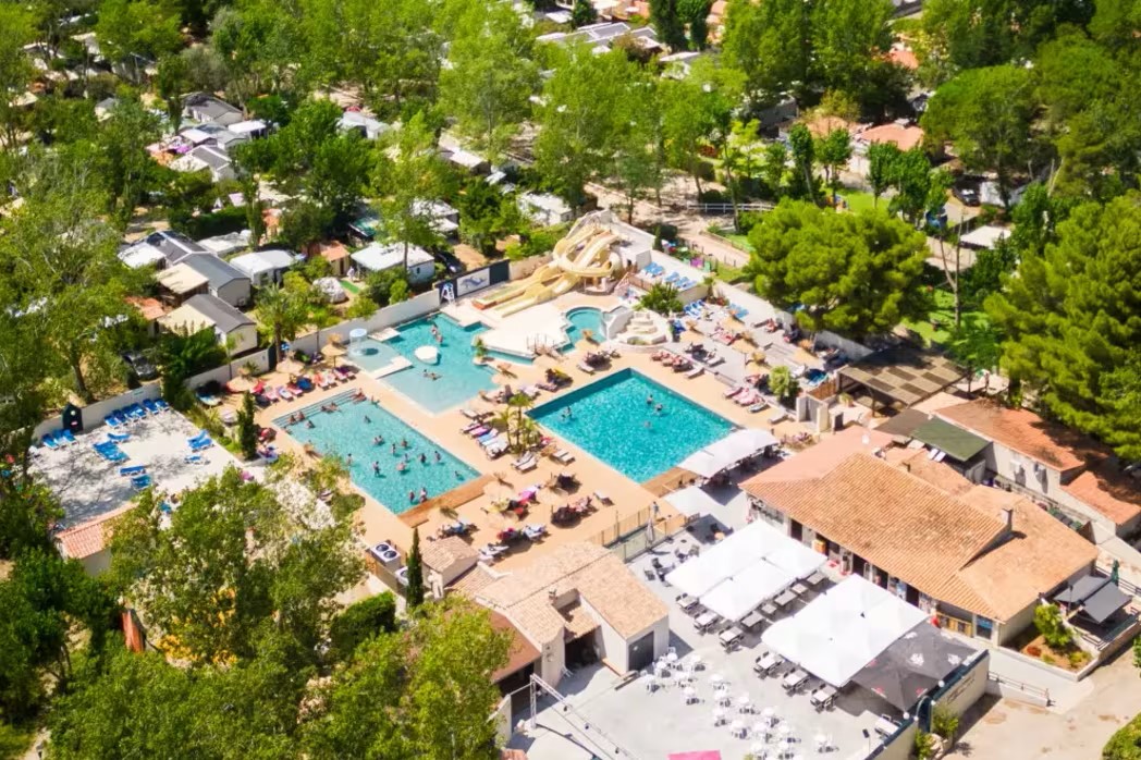 At Les Jardins de Tivoli, swimming pools and water slides teeming with individuals are encircled by lounge chairs and trees.