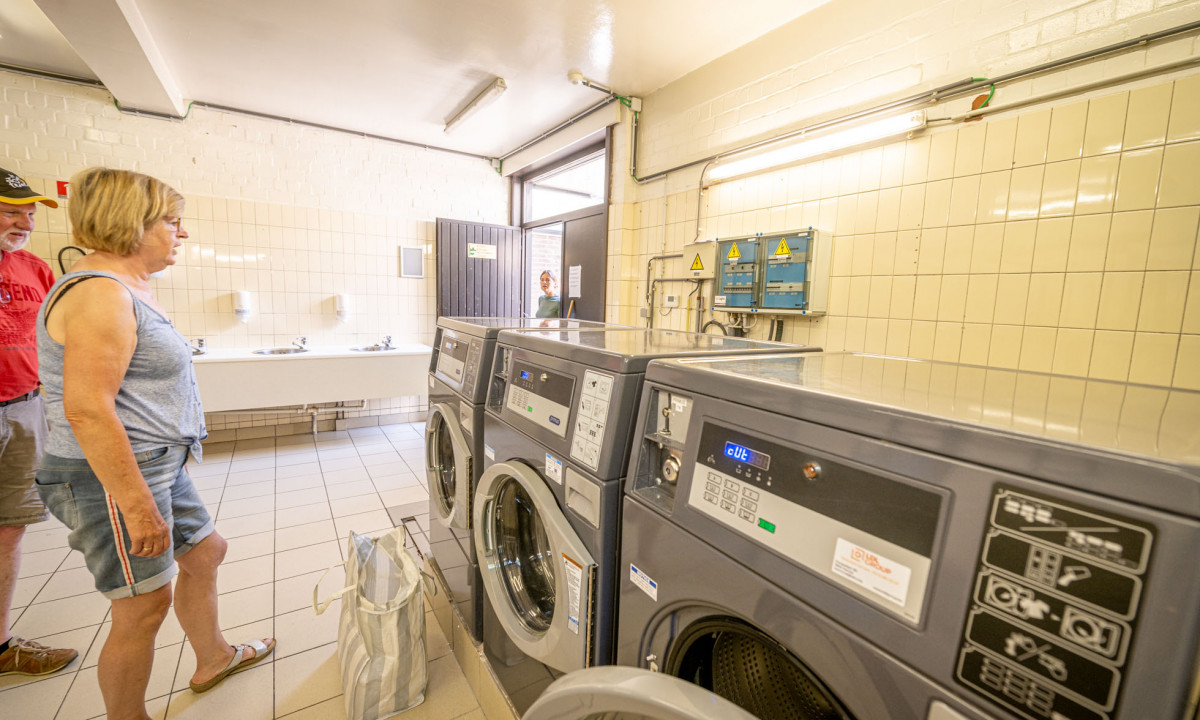 At Urban Gardens Gent, an individual is filling a washing machine in a well-lit, tiled laundry area, with other people present in the background.