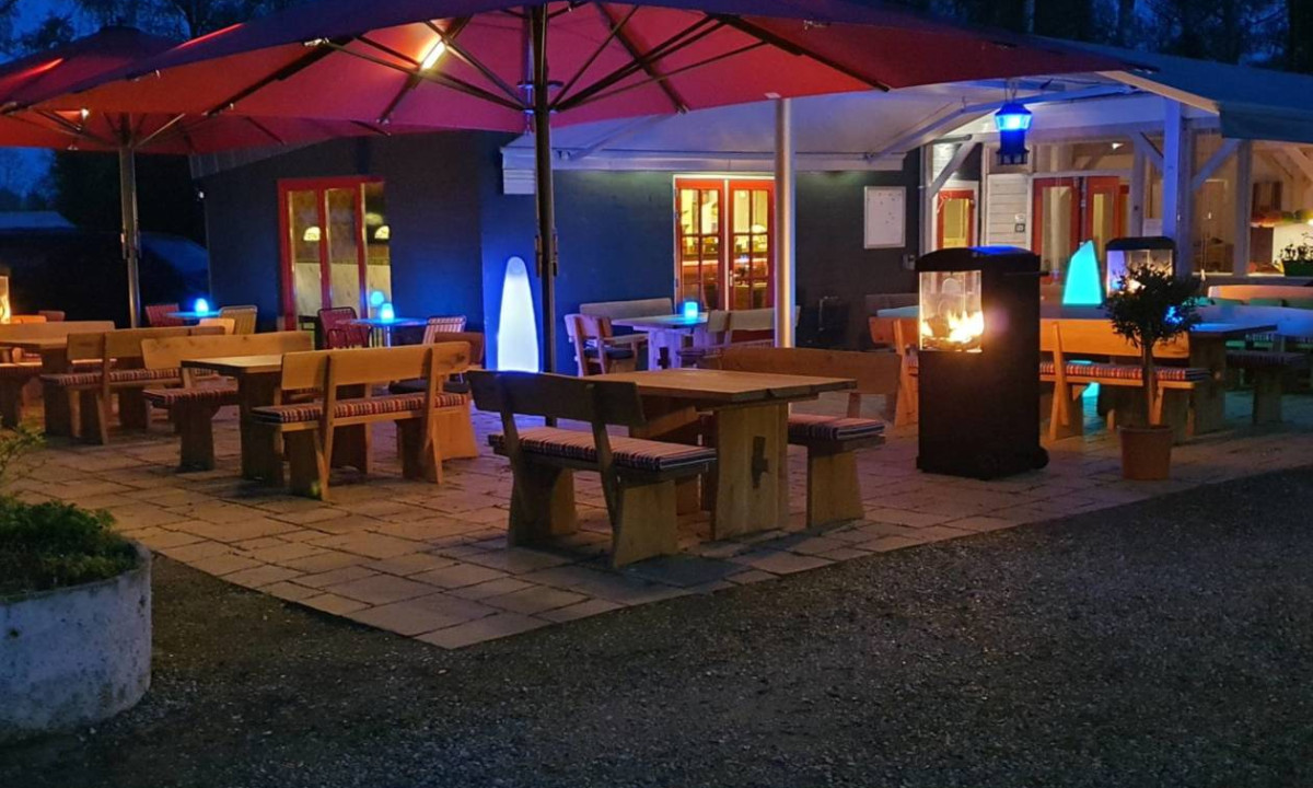 At Elbeling, wooden picnic tables and benches are situated beneath red umbrellas, lit by glowing lamps and a fire pit.
