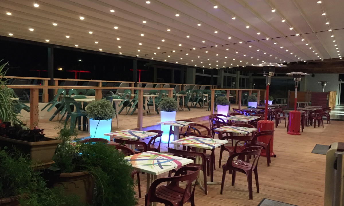 At La Clémentine, tables set beneath string lights are encircled by crimson chairs and planters on a wooden terrace.