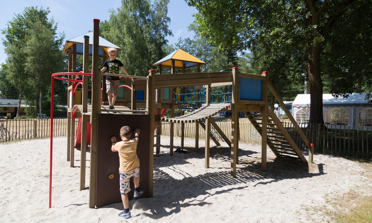 At het Veen, kids enjoy themselves on a timber play structure featuring slides and climbing zones, encircled by trees and tents.