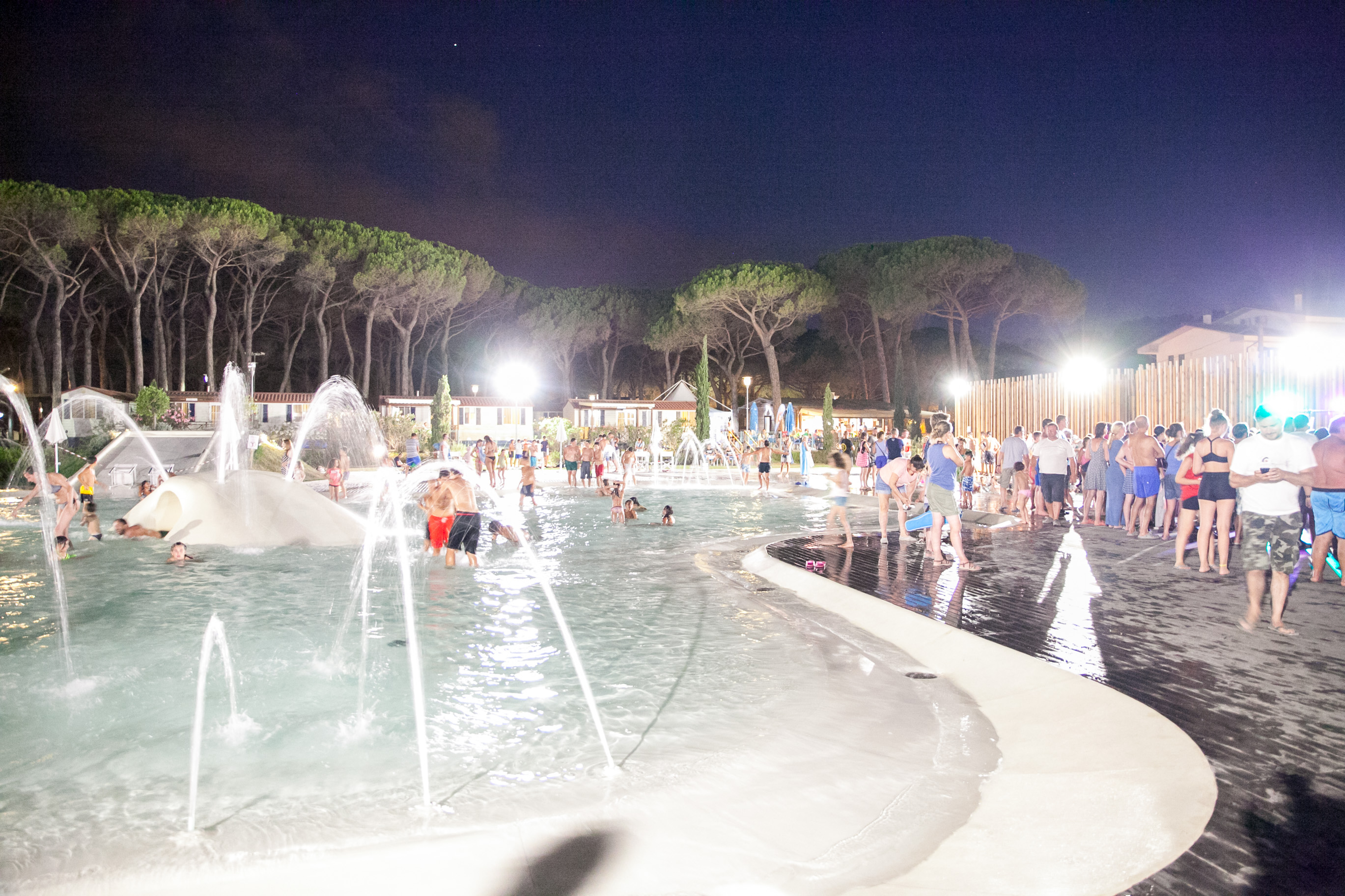 People swimming and playing in a lit outdoor pool surrounded by trees and vacation cabins at Pineta Sul Mare Camping Village