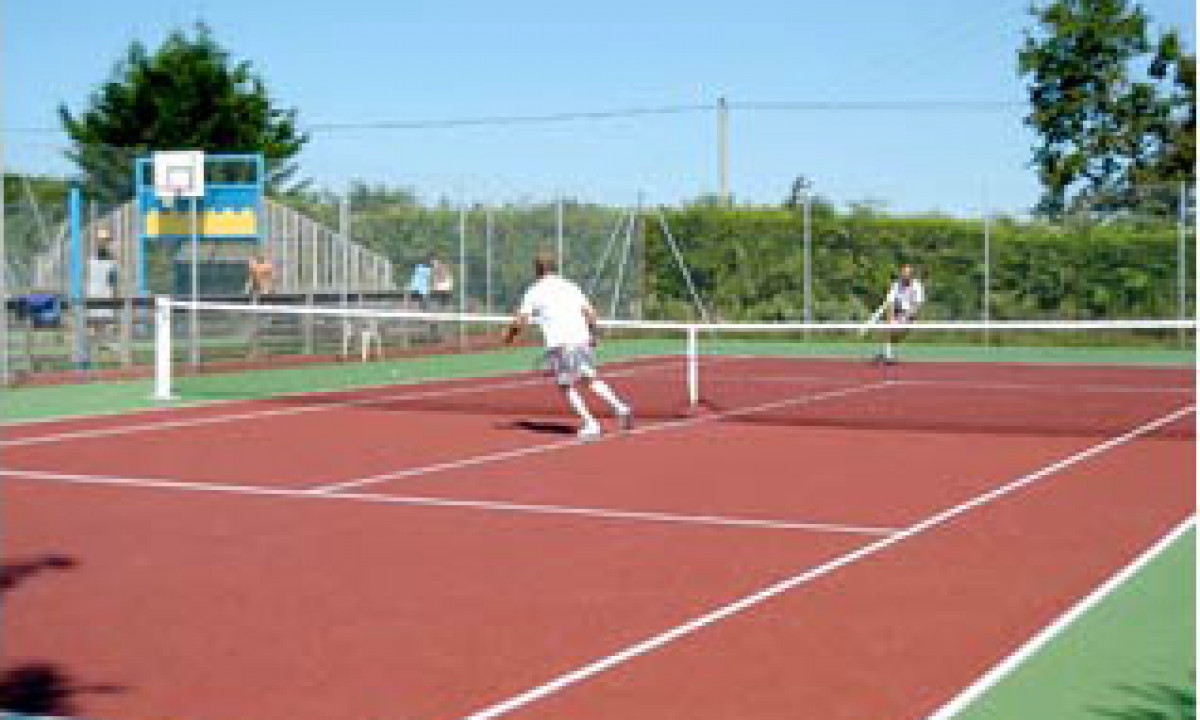 At Le Domaine de Léveno, two individuals engage in a game of tennis on an outdoor court encircled by trees and a basketball hoop.