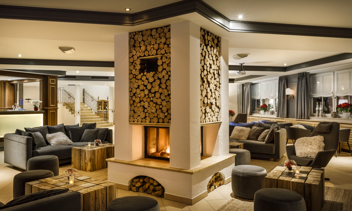 Central fireplace burning warmly, surrounded by cozy seating and wooden decor, within a spacious, elegantly furnished lounge at Valamar Obertauern Hotel