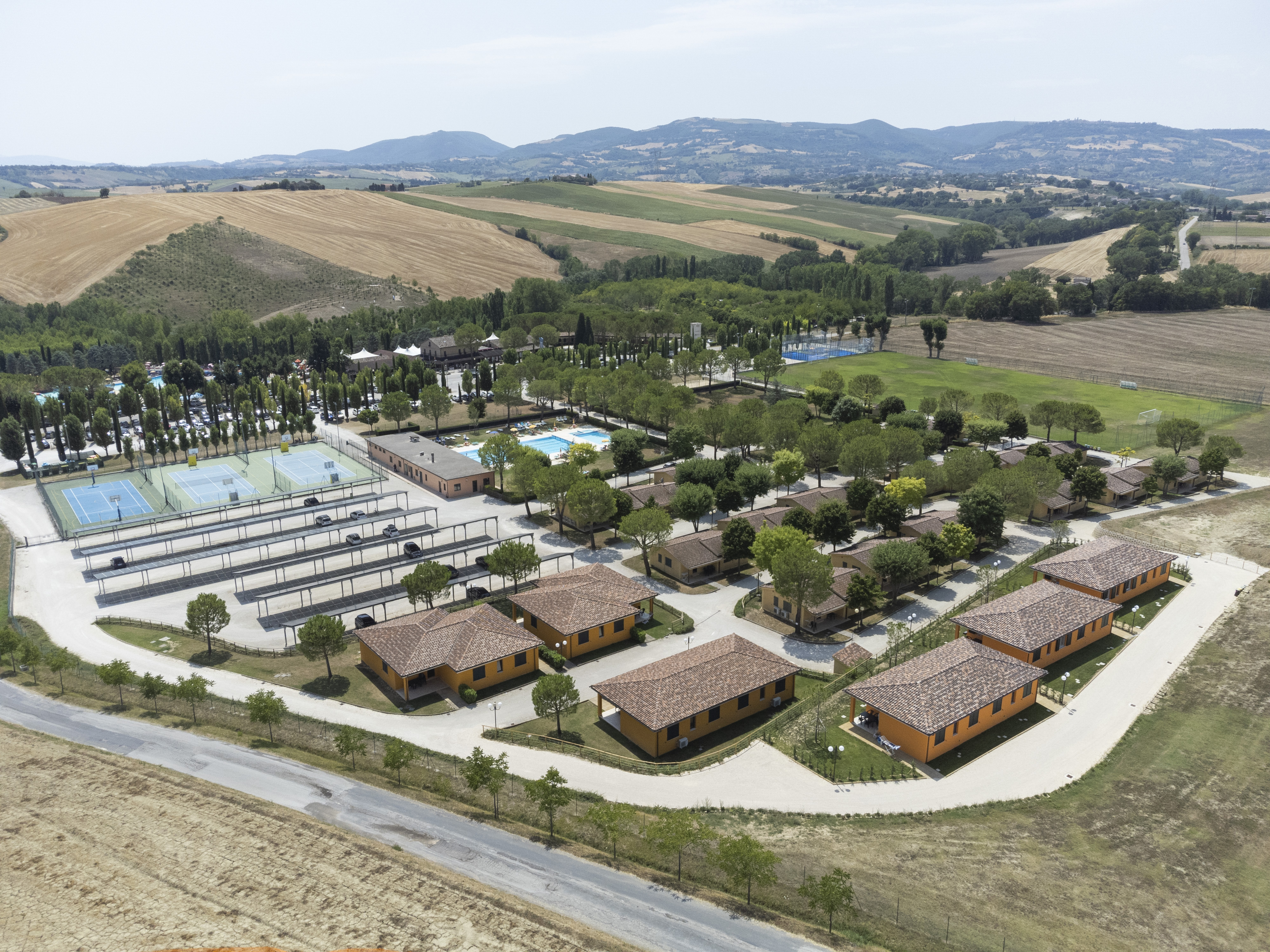 Aerial view of cabins arranged around sports facilities, surrounded by fields and distant mountains and hills at Centro Vacanze Verde Azzurro