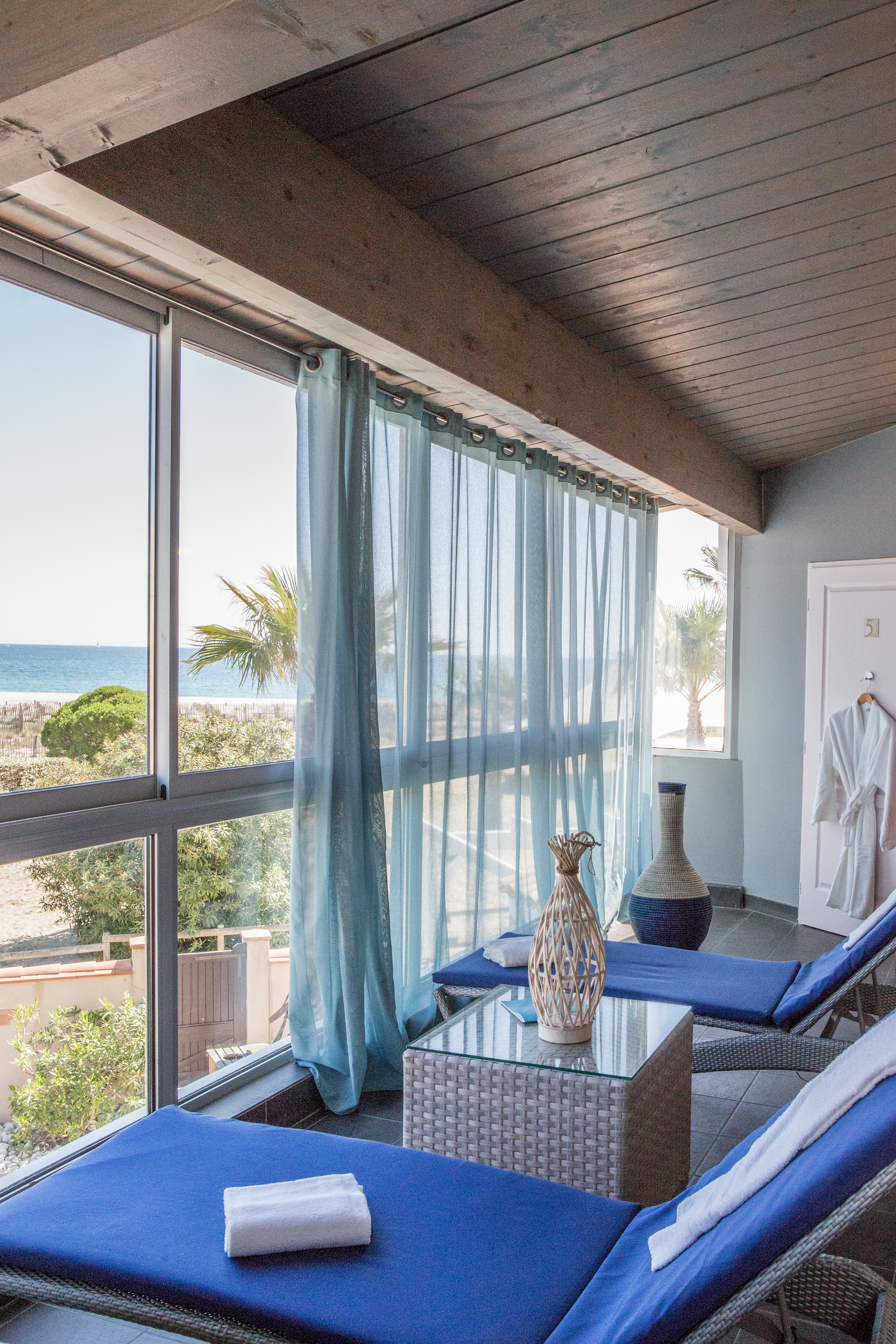 Two lounge chairs with blue cushions face a large window overlooking a beach, adorned with light blue curtains at Les Bulles de Mer