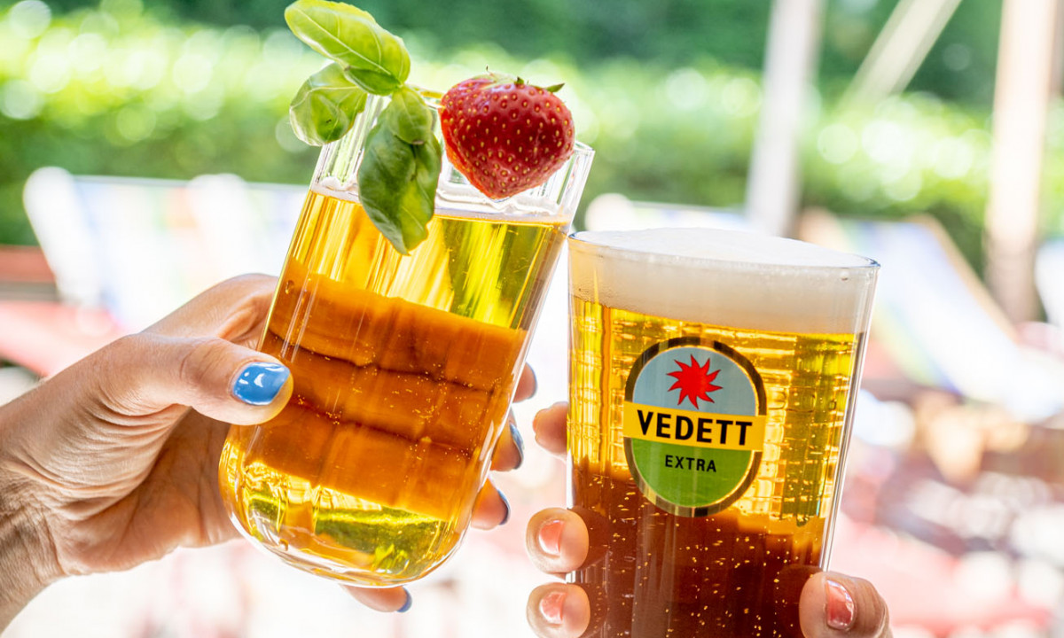At Urban Gardens Gent, two hands clinking beer glasses together outdoors beneath a blue umbrella.