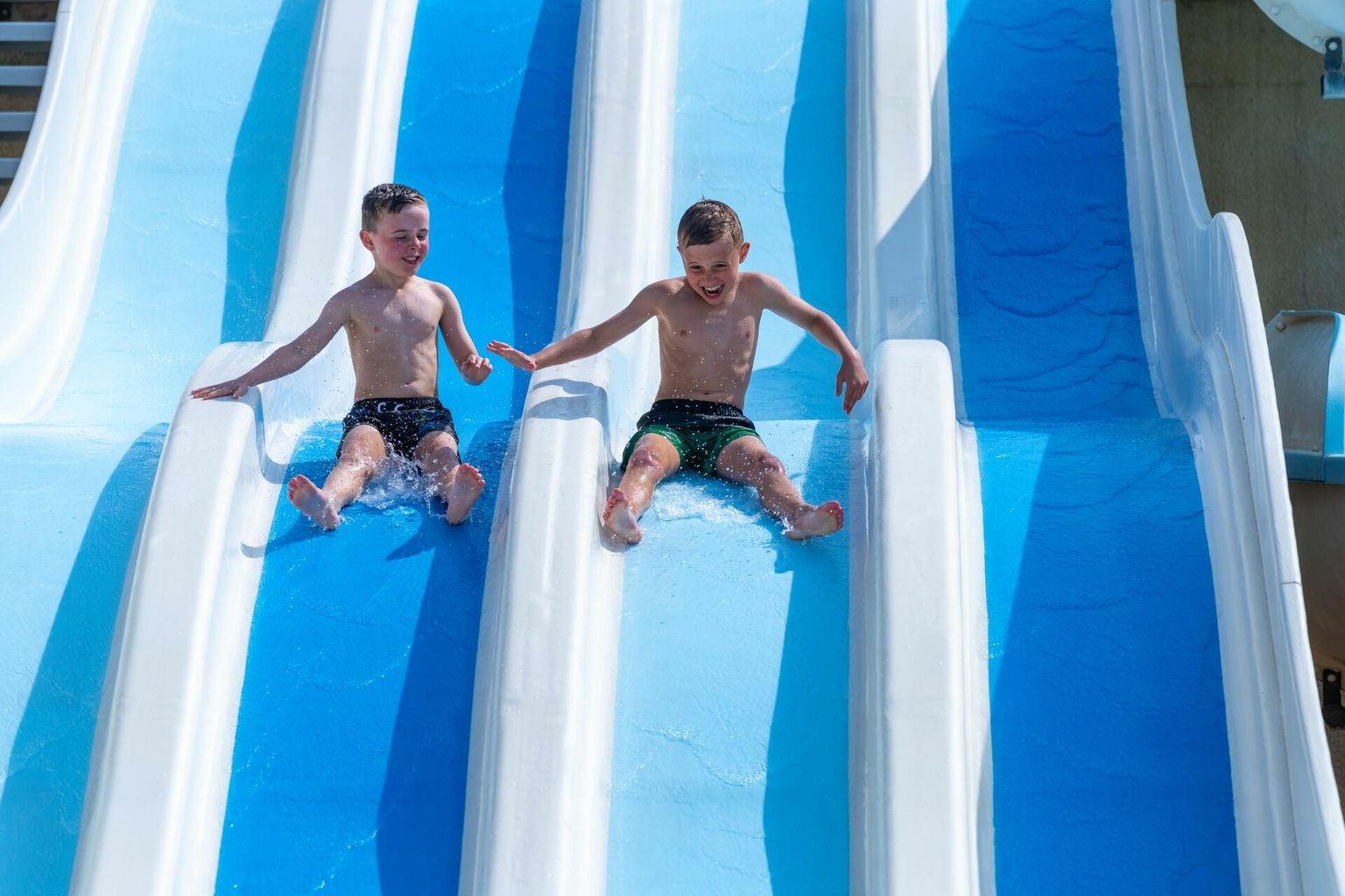 At Le Carbonnier, two kids glide down neighboring blue water slides, giggling in a bright outdoor environment.