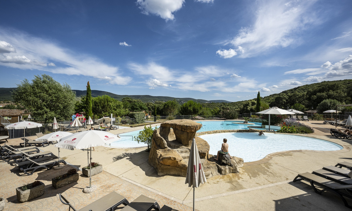 Pool with rock feature and sunbathers under umbrellas, surrounded by lush greenery and mountains at Domaine de Sévenier