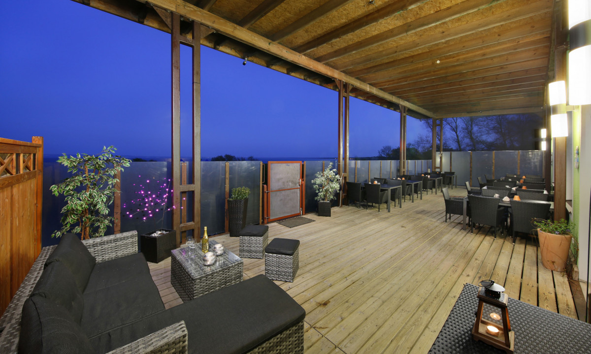 At KNAUS Campingpark Rügen, an outdoor seating space showcases wicker furnishings and greenery on a timber deck, aglow at dusk.