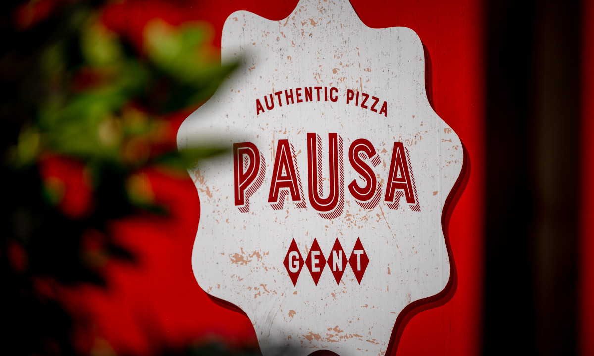 At Urban Gardens Gent, a sign displaying "Pausa Authentic Pizza Gent" is set against a red backdrop, partially hidden by blurred green vegetation.