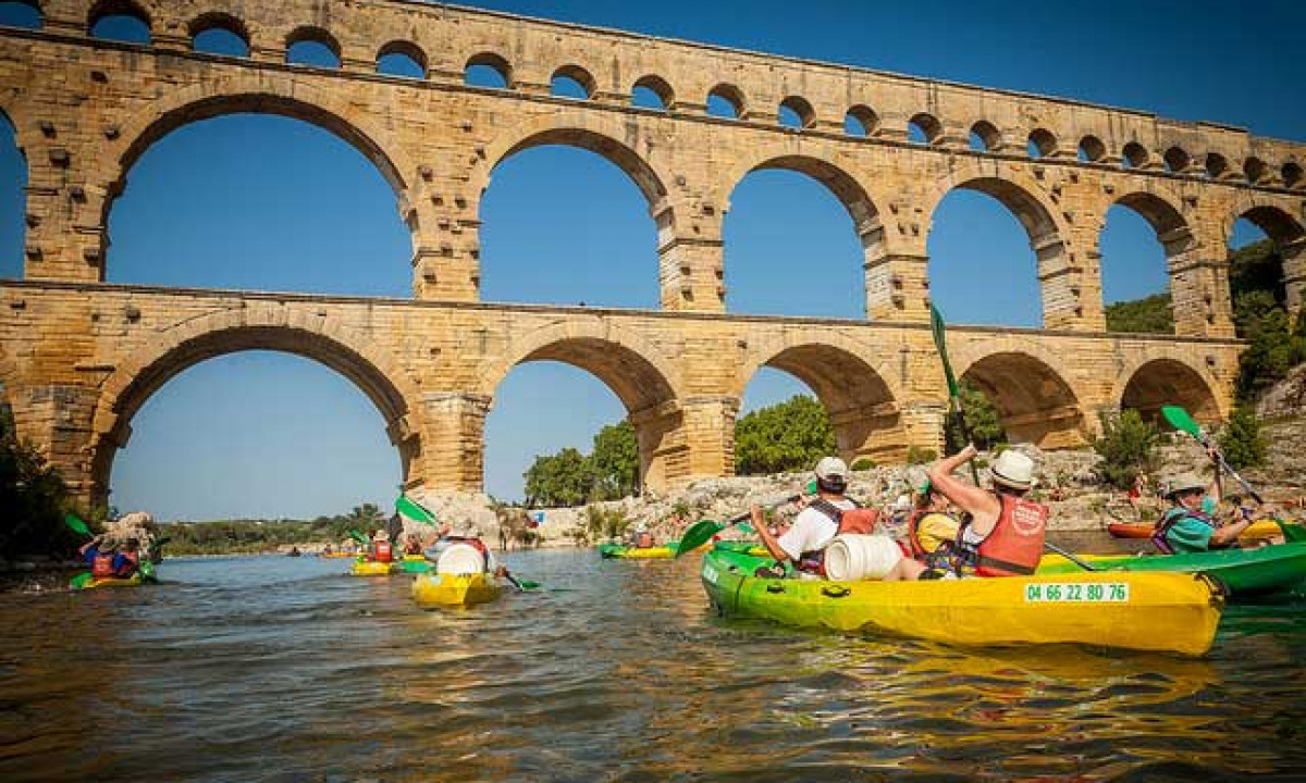 At La Sousta, kayakers are paddling beneath a historic stone aqueduct on a bright day, surrounded by lush vegetation along the riverbanks.