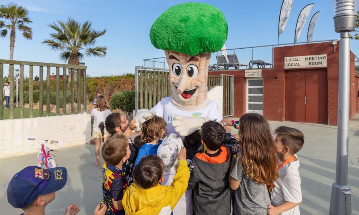 Broccoli mascot entertaining a group of children outdoors near a building labeled "Local Social Meeting Room" at Resort Els Pins
