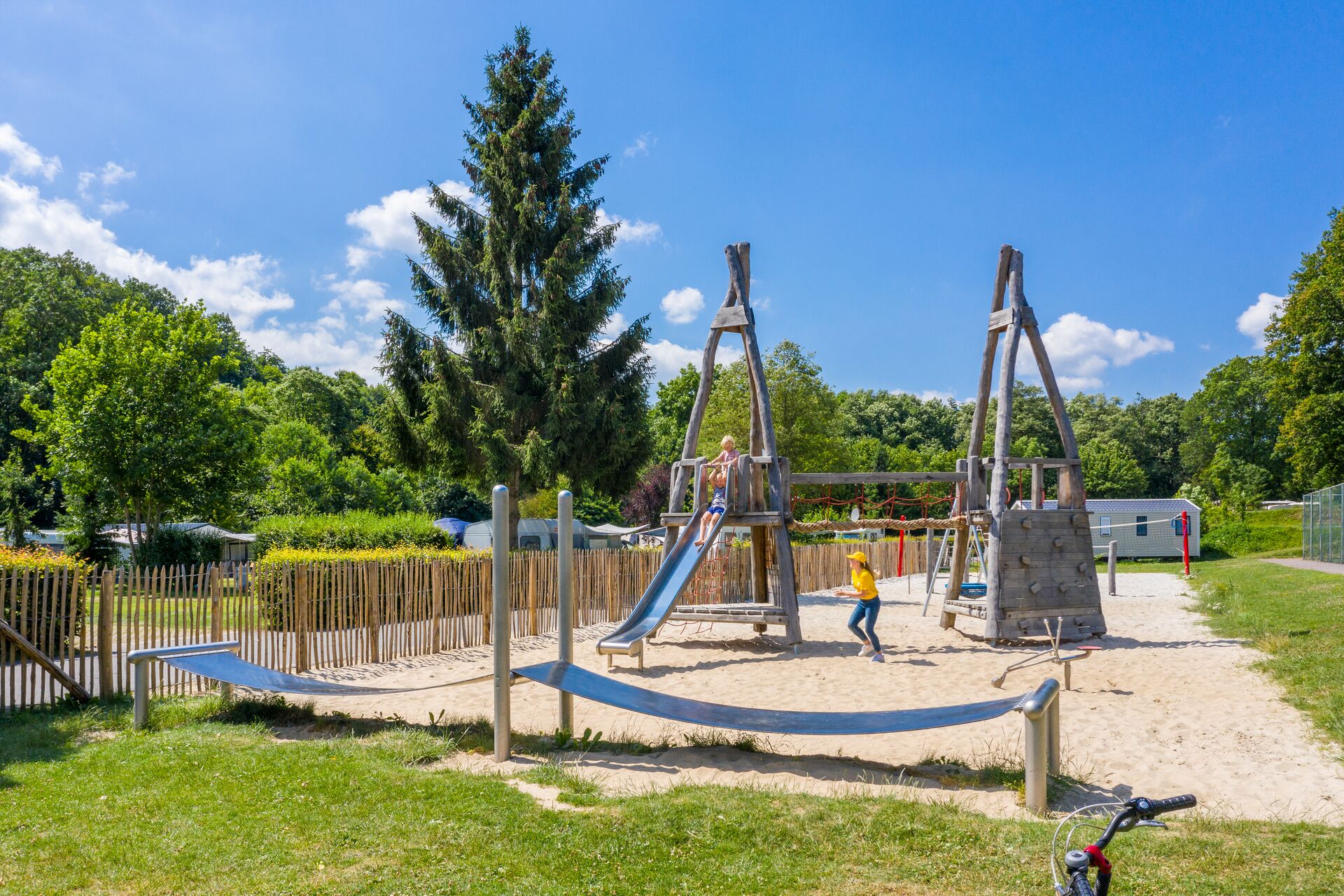At Parc La Clusure, kids are enjoying themselves in a wooden play area featuring a slide and climbing equipment, encircled by trees and grass.