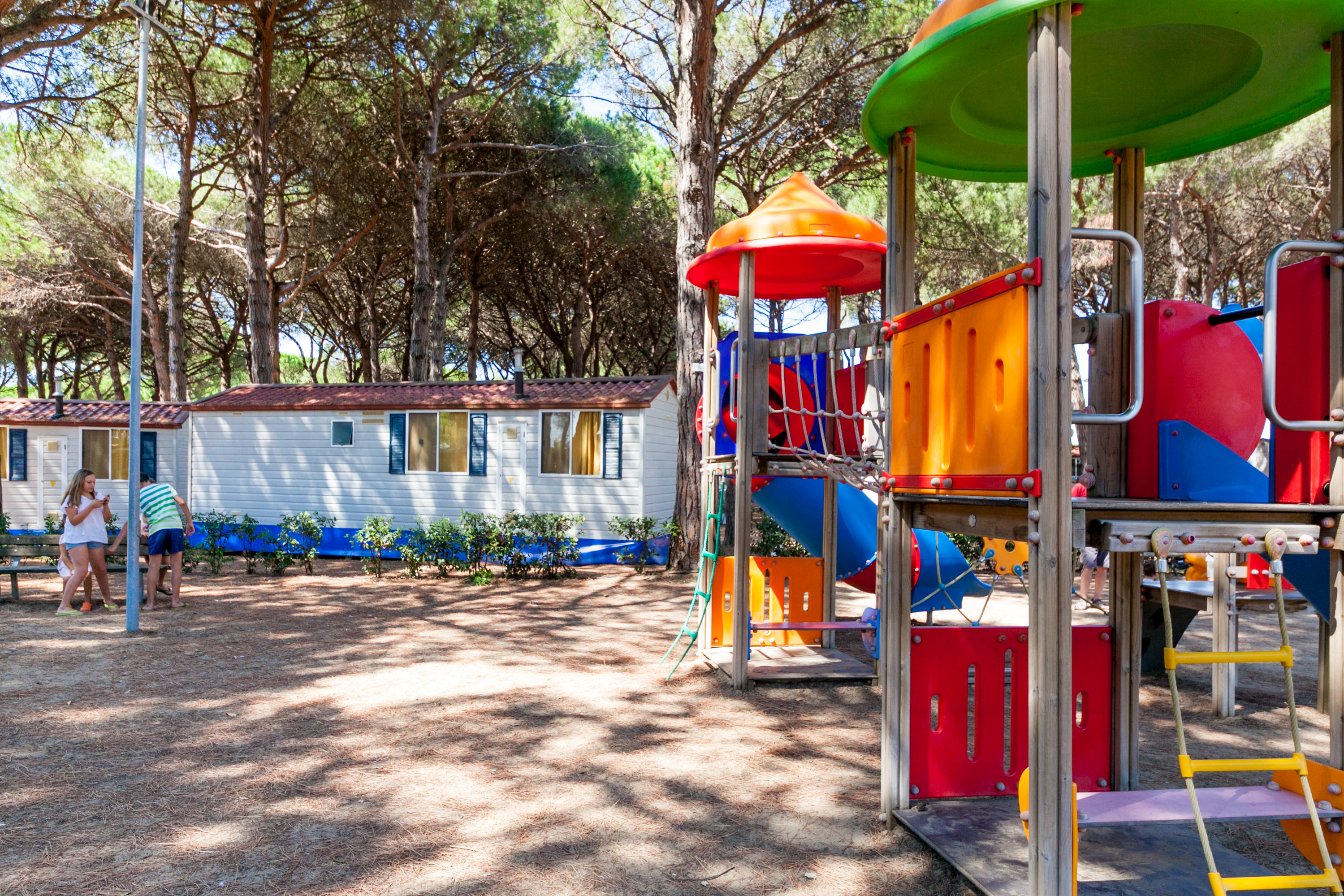 Playground equipment standing still, bright colors, surrounded by cabins and trees. People nearby under the shade of tall trees at Pineta Sul Mare Camping Village