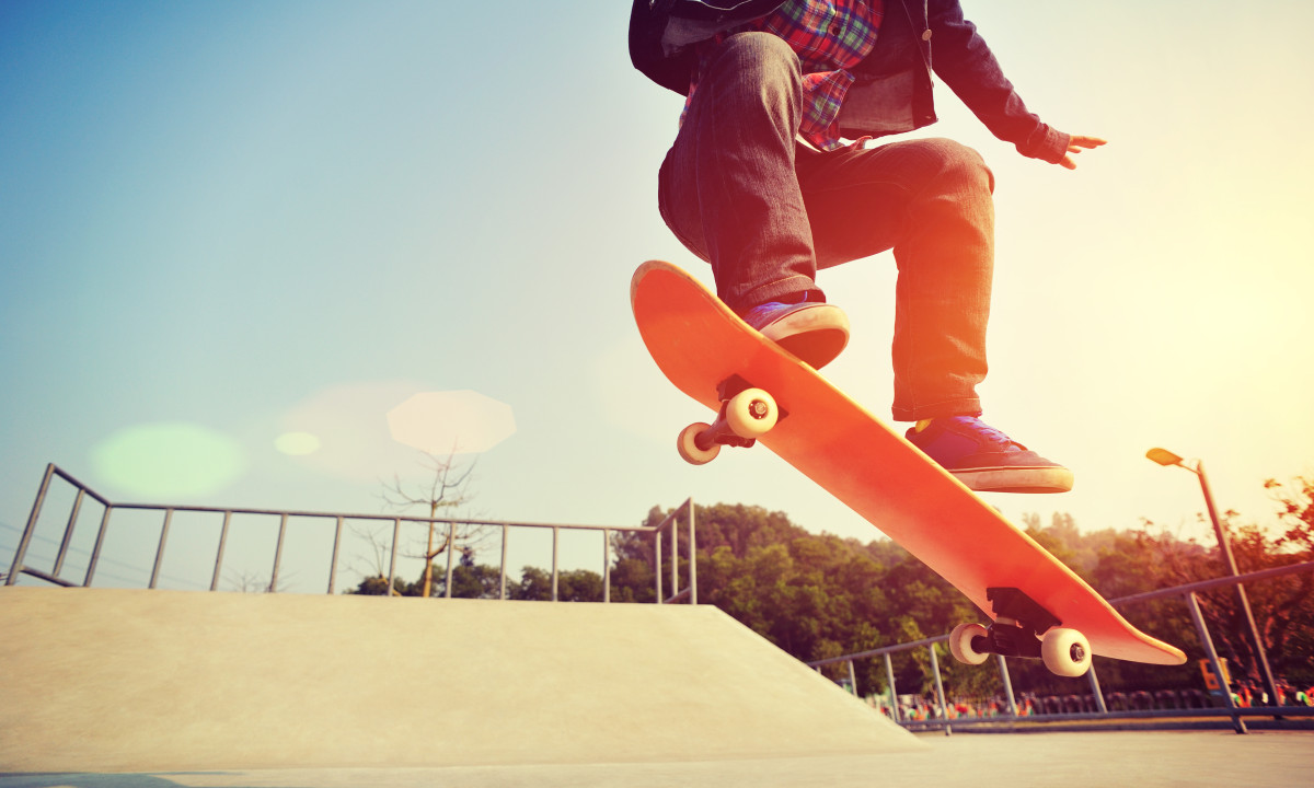 At Urban Gardens Gent, a skateboarder is executing an aerial maneuver on a ramp in a bright outdoor skate park, with trees in the backdrop.