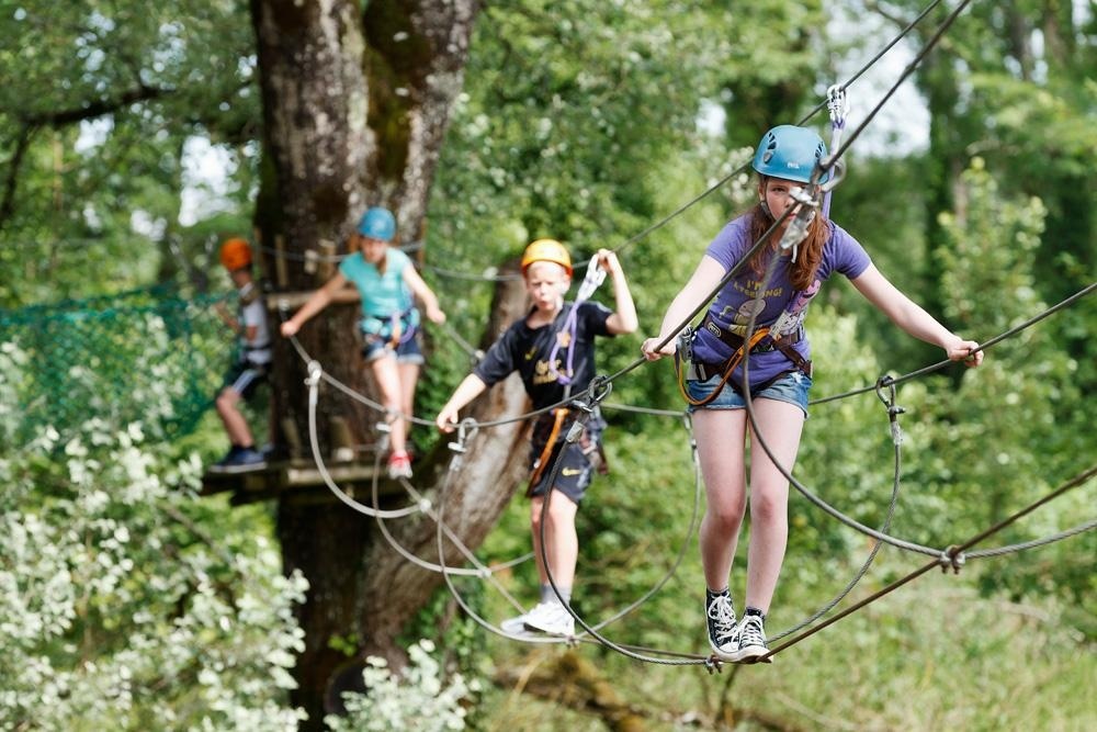 Children with helmets balancing on a high rope course in a wooded area, using harnesses for safety at Le Val de Bonnal