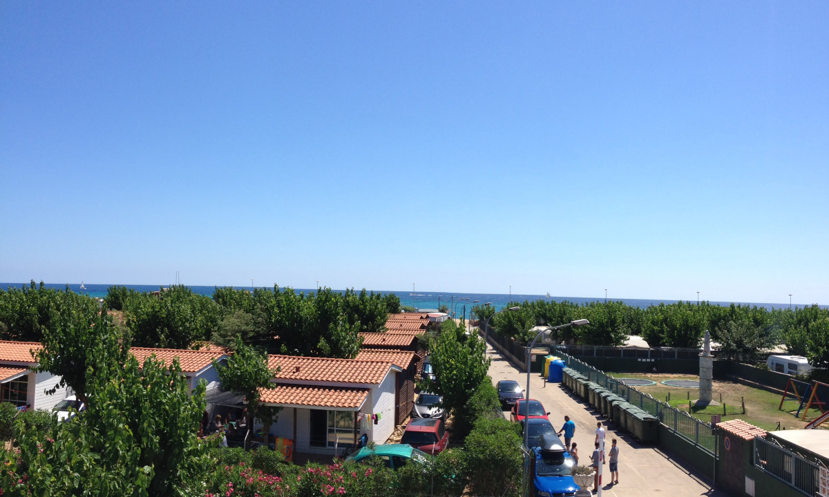 Rows of small houses with red roofs, surrounded by trees, leading to the beach under a clear blue sky at Resort Els Pins