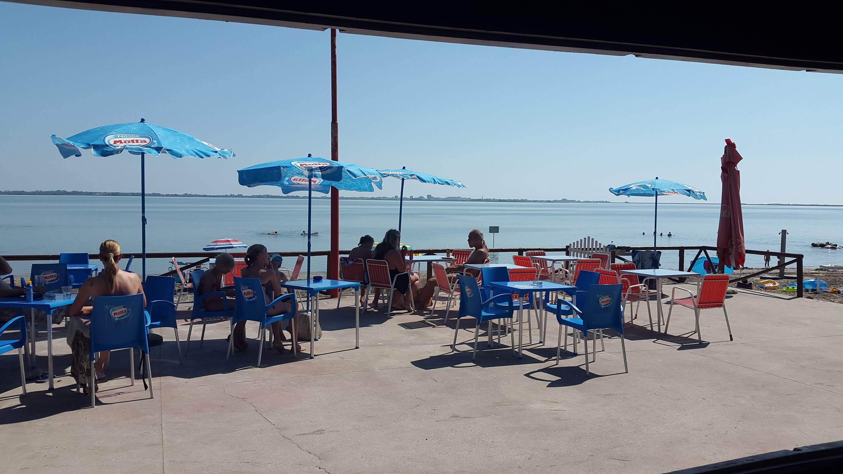 People sit under blue umbrellas at outdoor seaside café, a calm ocean and horizon visible in the background at Belvedere Pineta