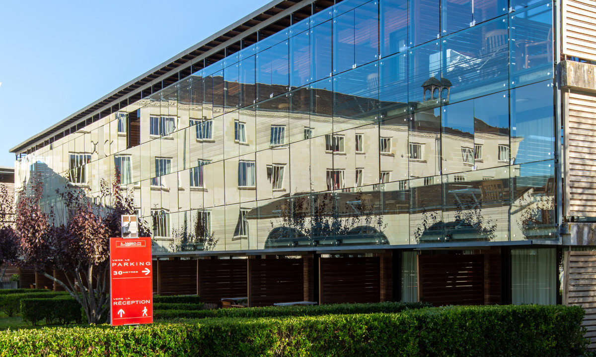 A glass-walled building reflecting another building, surrounded by green hedges and trees in a sunny outdoor setting at Le Moulin des Cordeliers