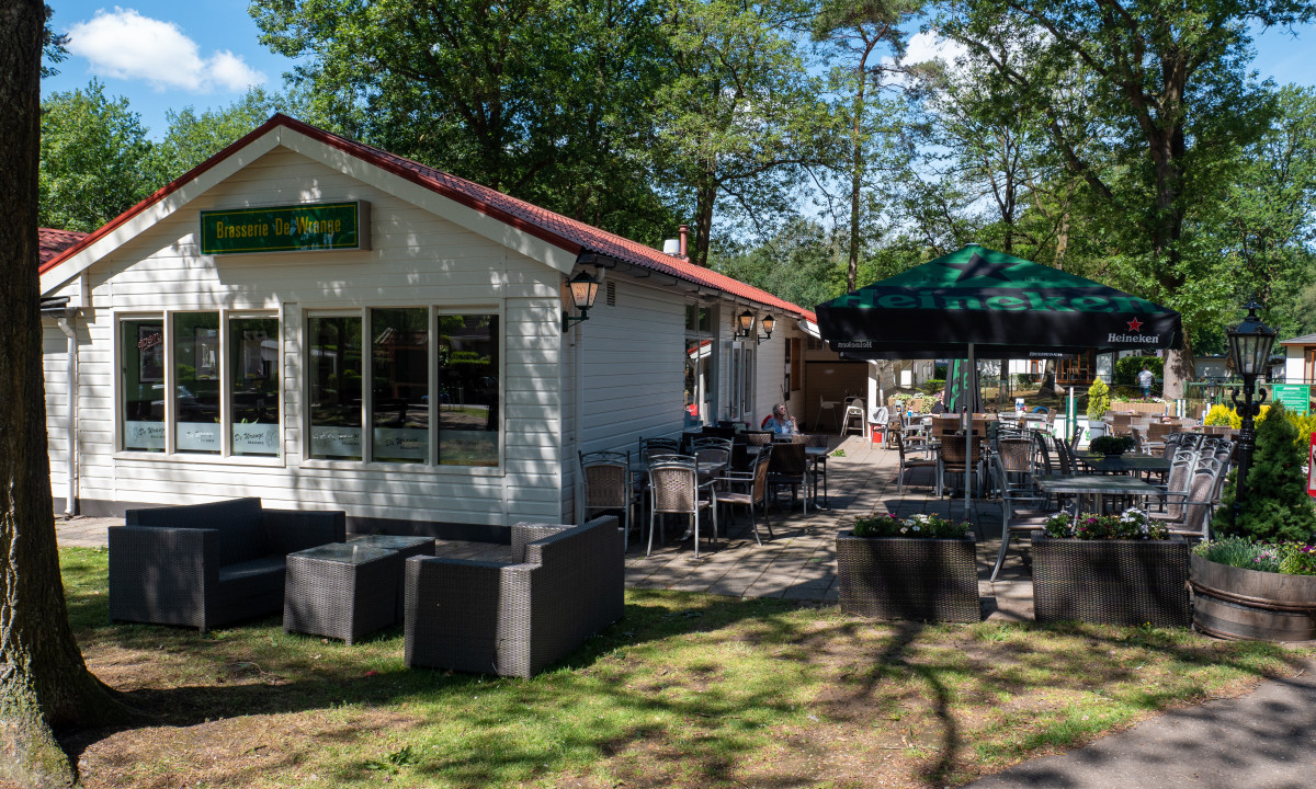 Outdoor café with tables, chairs, and a large umbrella, set near a white building with a red roof among trees at de Wrange