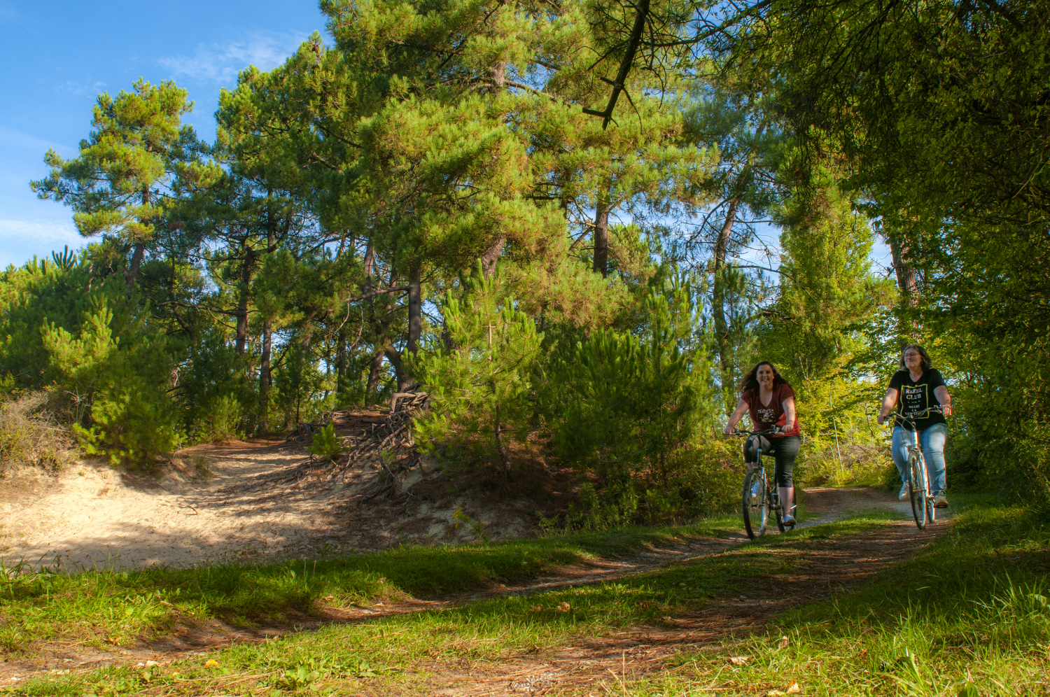 At La Dune Blanche, two bikers pedal along a forested trail, encircled by vibrant green trees on a bright day.