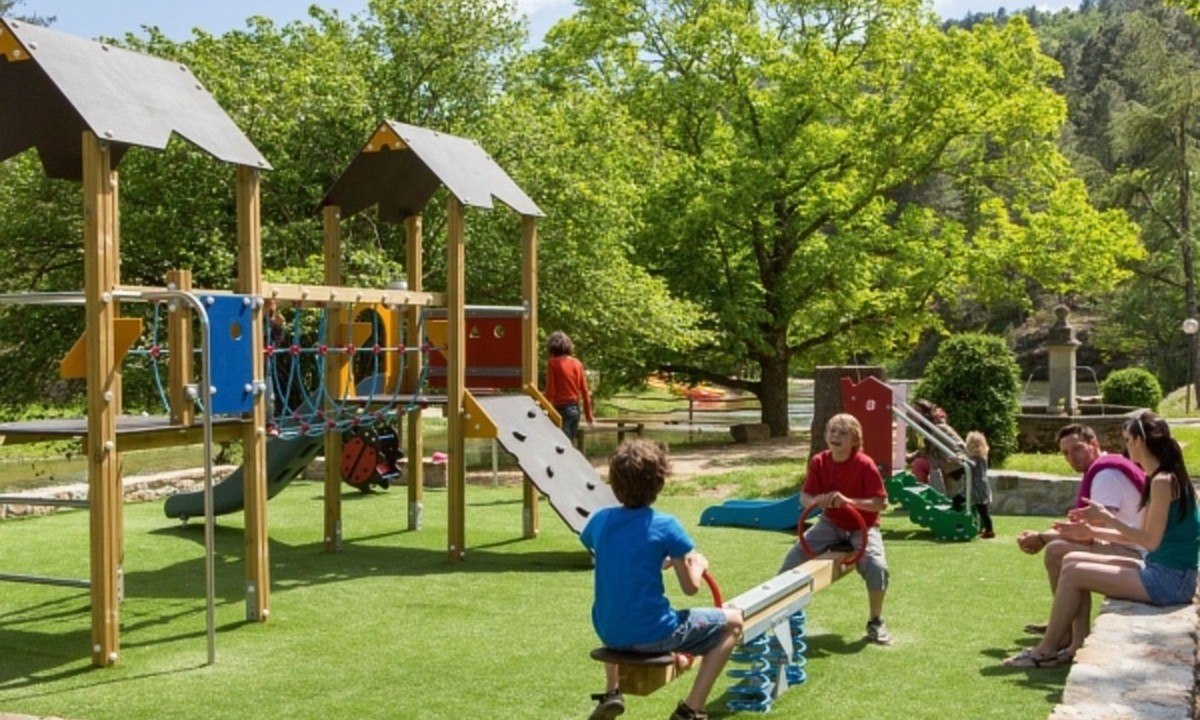 At Les Ranchisses, youngsters are enjoying themselves on a play structure featuring a slide and seesaw, encircled by vibrant trees and adults conversing.