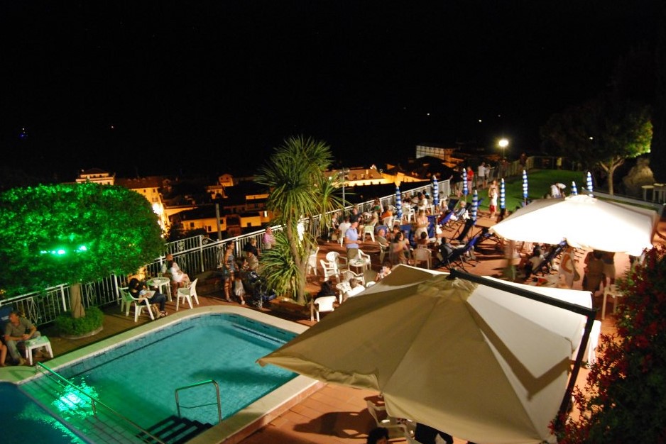 A large group of people is dining and mingling by a pool under the night sky, atop a terrace overlooking a town at Sant' Anna Residence