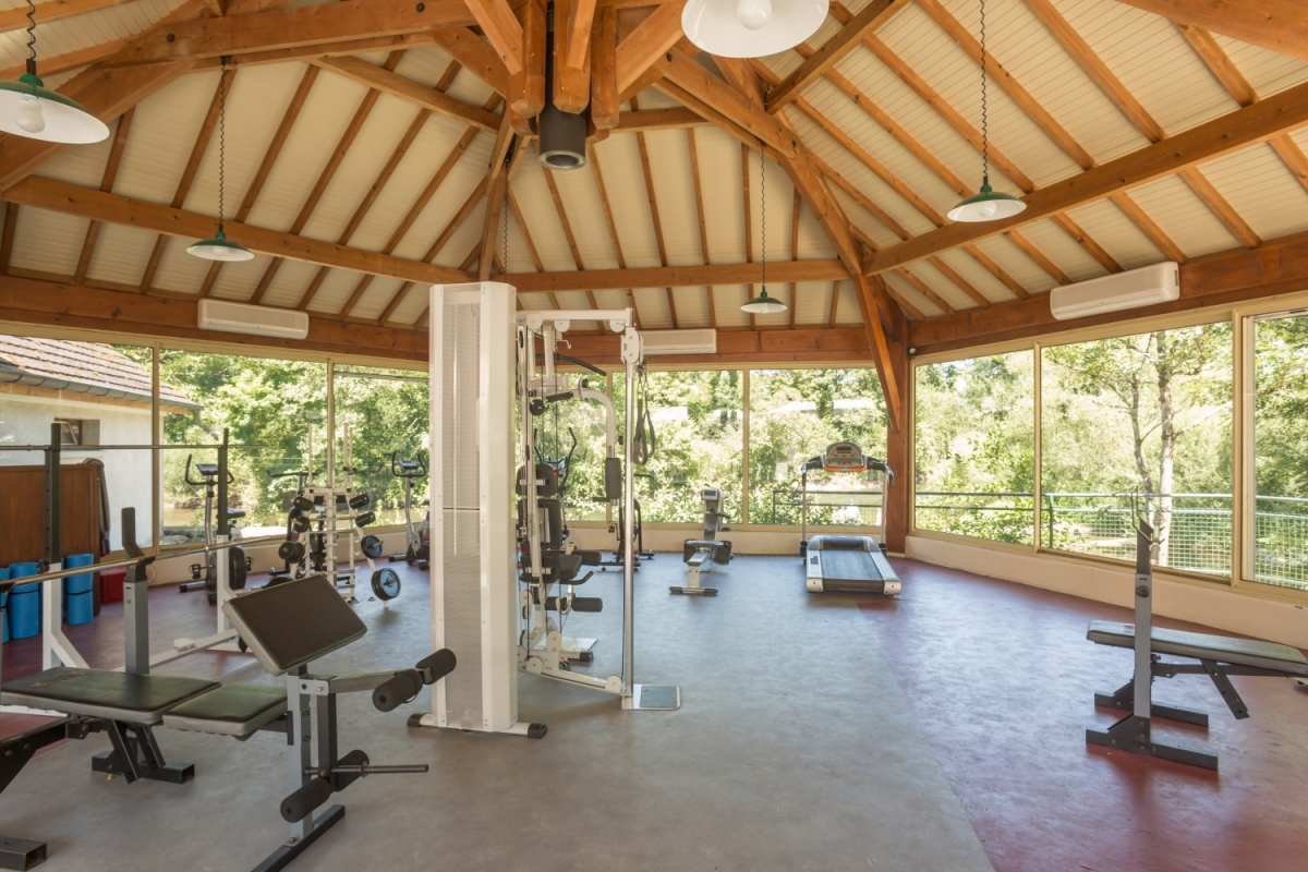 A gym, featuring various exercise machines, is situated under a wooden pavilion with large windows overlooking a leafy environment at Le Val de Bonnal